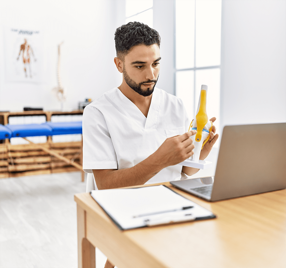 Young man wearing physiotherapist uniform having video call holding anatomical model of knee